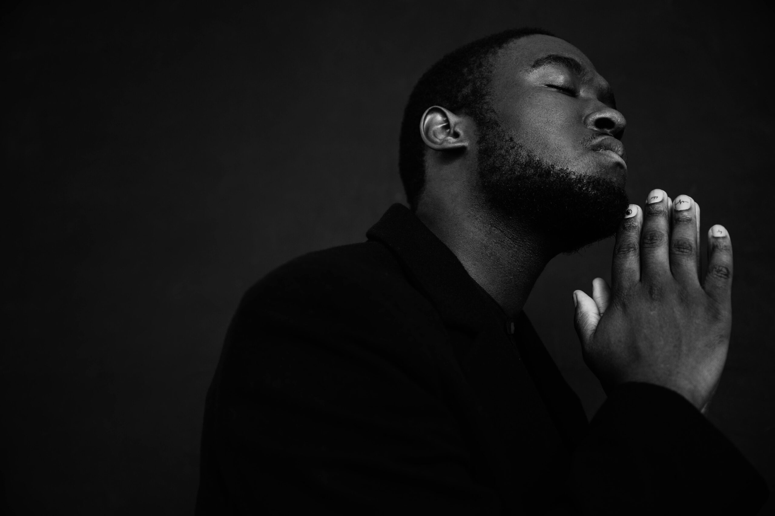Monochrome handsome unshaven African American male in black suit praying with hands clasped under chin