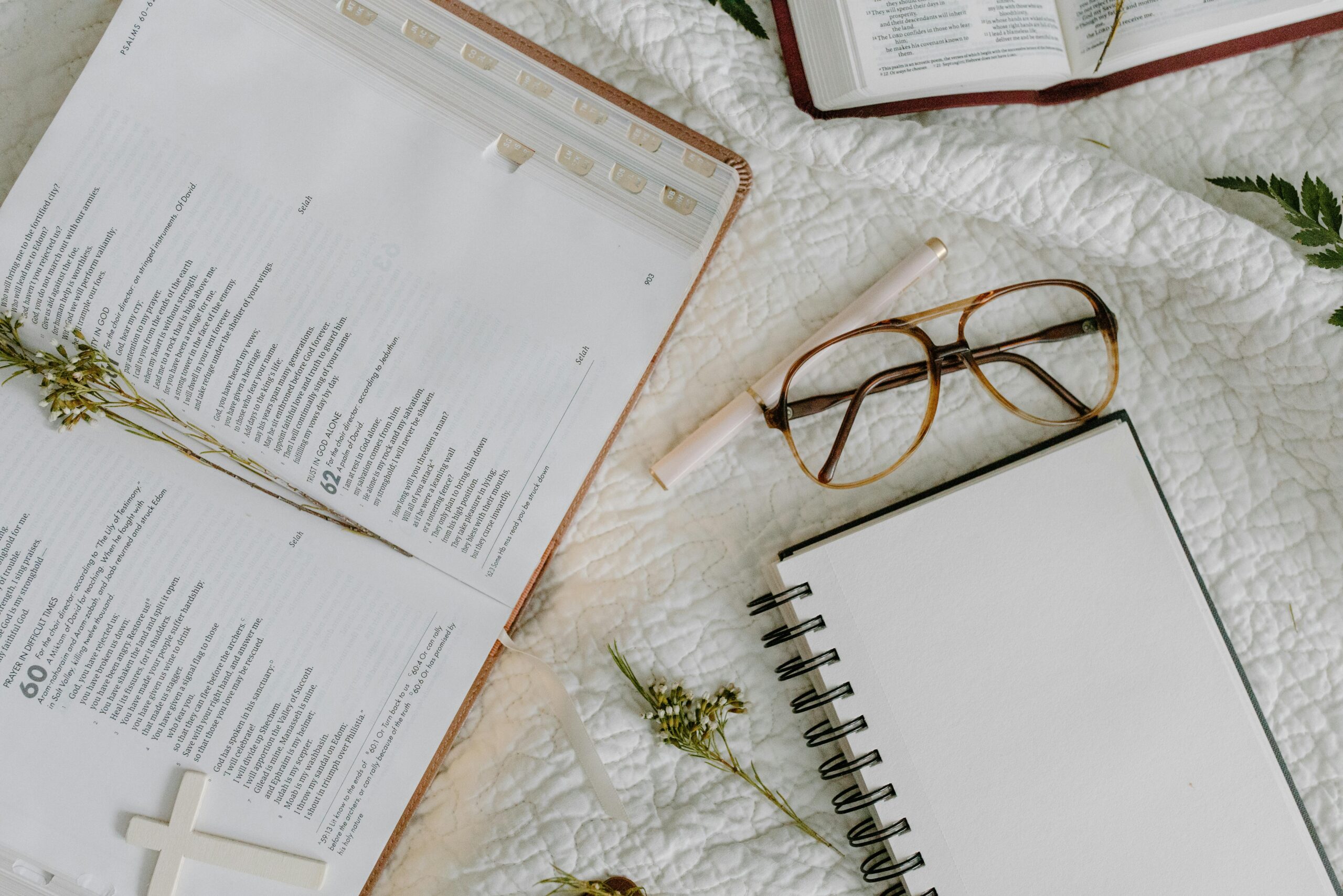 An open Bible with glasses and a blank notebook on a cozy bedspread setting.